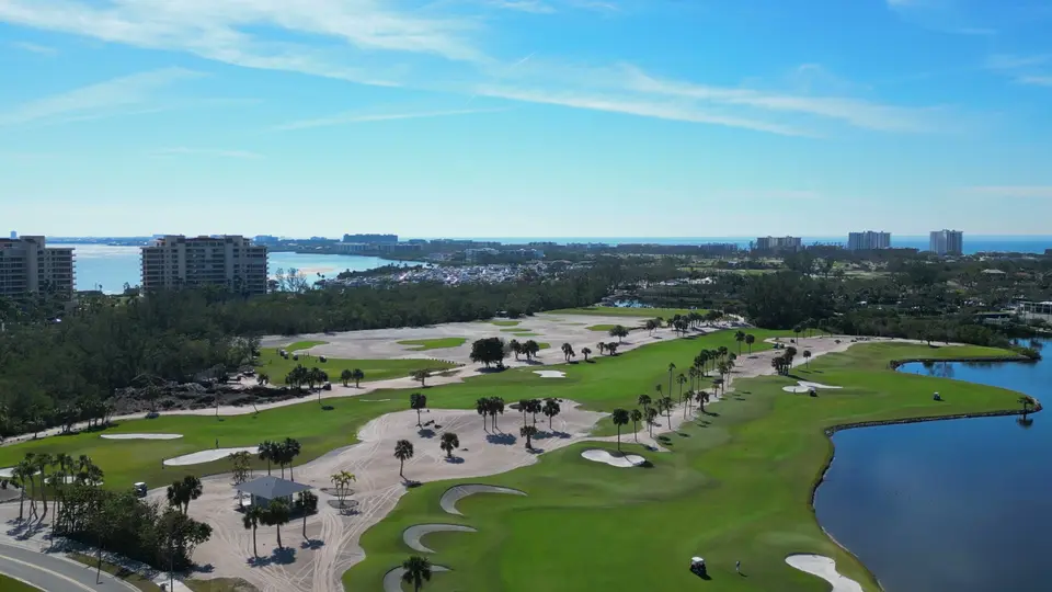 Aerial of golf course greens and a lake with Gulf-front towers beyond