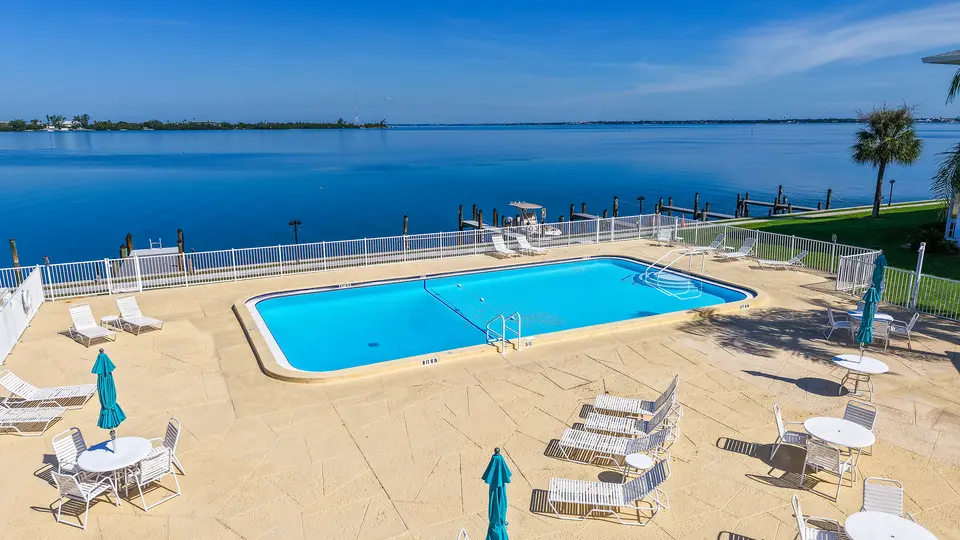 Community pool deck with lounge chairs beside open bay water under a clear sky